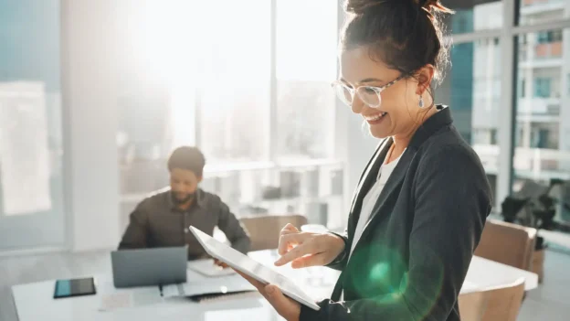 In a modern office environment, a smiling woman looks down at a tablet.
