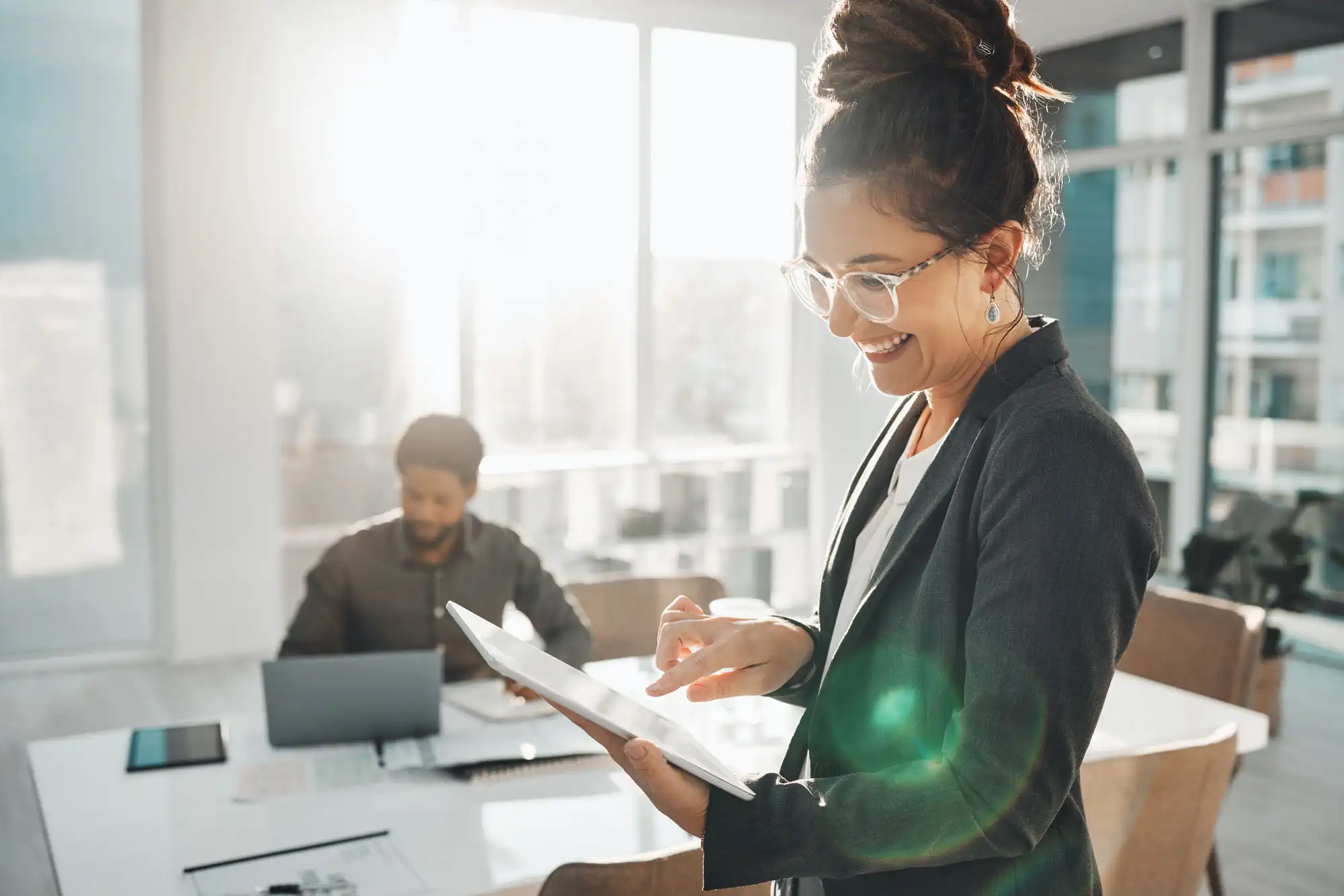 In a modern office environment, a smiling woman looks down at a tablet.
