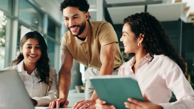 Three smiling office employees gathered around a laptop, at a desk.