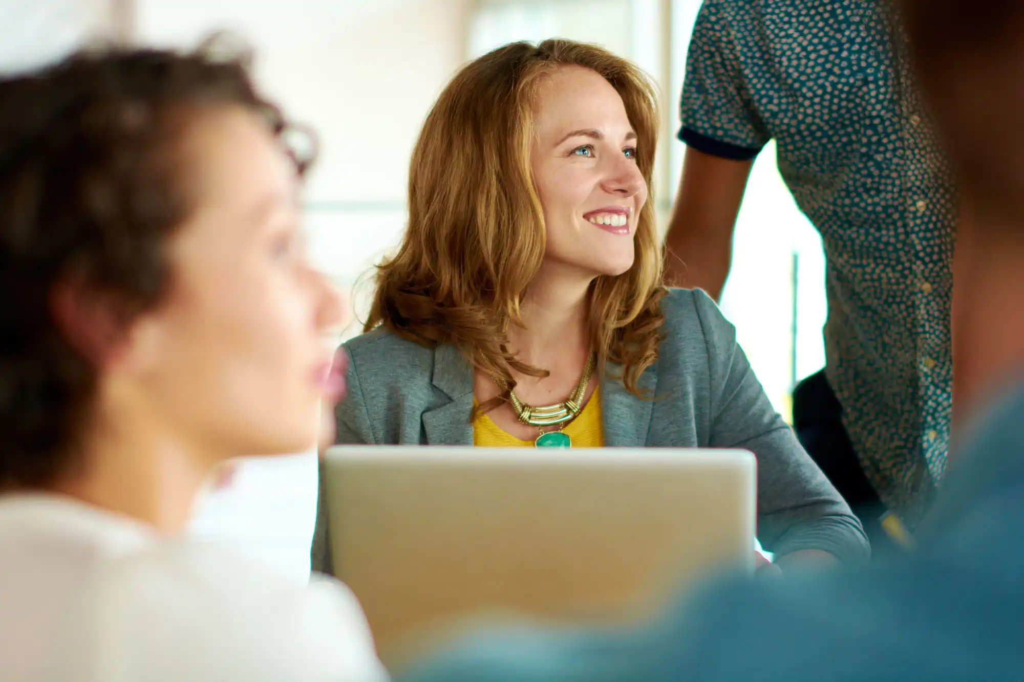 A businesswoman smiles in front of her laptop, while listening to someone offscreen.