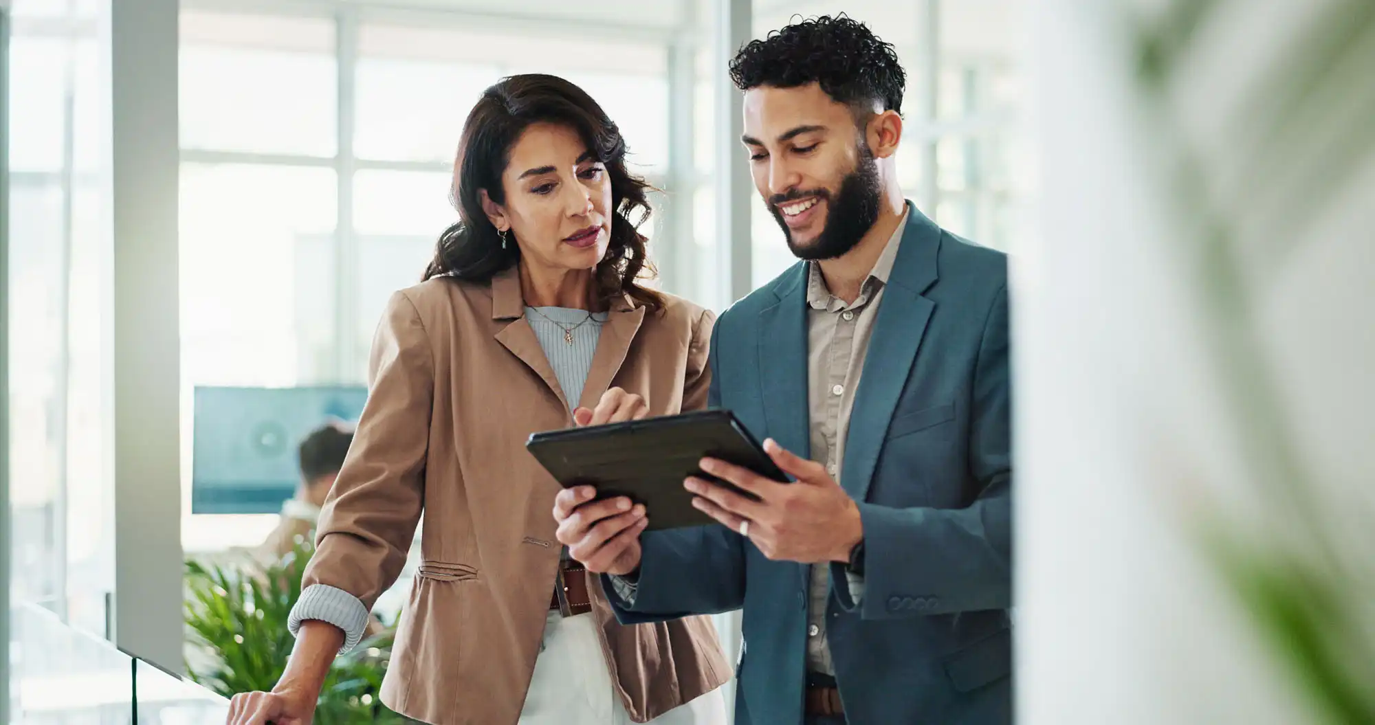Two business professionals in an office setting, reviewing information on a tablet.